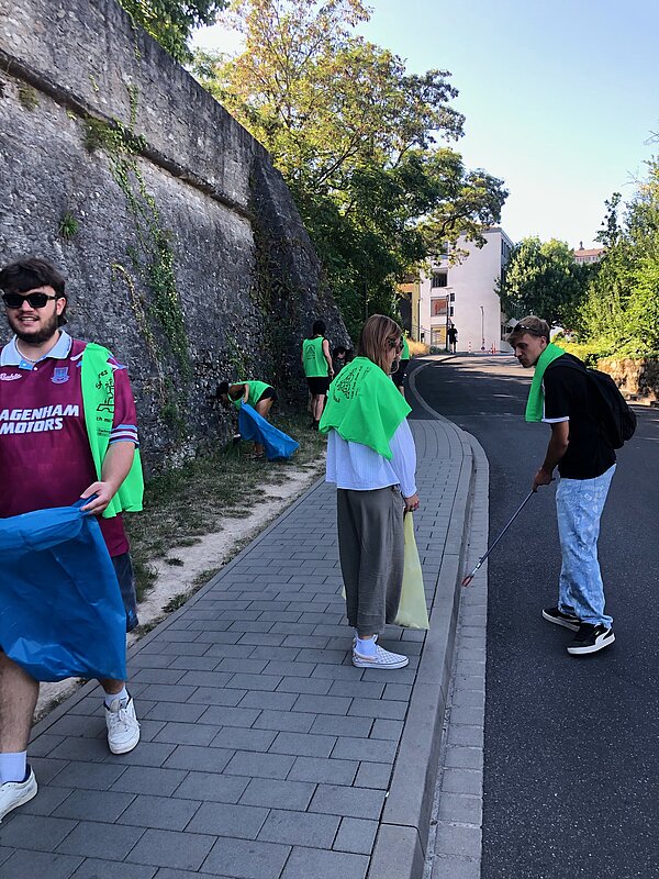 Eine Gruppe von Personen in grünen Westen sammelt Müll auf einem Gehweg. Einige Personen tragen blaue Müllsäcke.