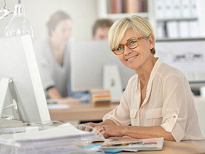 Eine ältere Frau mit kurzen, hellen Haaren und einer Brille sitzt lächelnd an einem Schreibtisch in einem Büro. Sie trägt ein hellbeiges Oberteil und arbeitet an einem Computer. Auf dem Schreibtisch liegen verschiedene Unterlagen, ein Tablet und ein Stift. Im Hintergrund sind unscharf zwei Kollegen zu sehen, die ebenfalls an ihren Arbeitsplätzen sitzen. Der Raum wirkt hell und freundlich, mit Regalen und Ordnern im Hintergrund. Die Frau wirkt zufrieden und konzentriert.