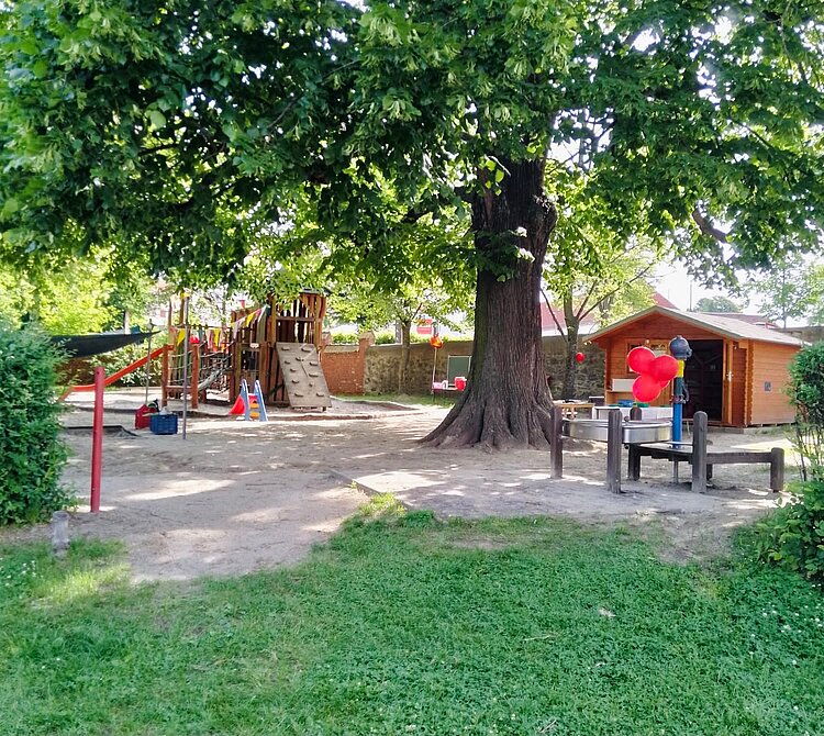 Ein Spielplatz in einem grünen, schattigen Gartenbereich. Im Vordergrund steht ein großer Baum mit dichtem Blätterdach. Links sieht man Spielgeräte wie eine Rutsche, ein Klettergerüst und eine Schaukel. Rechts befindet sich ein kleines Holzhaus mit roten Ballons und einem Wasserbecken. Der Boden ist mit Sand bedeckt, und im Hintergrund sind weitere Bäume und Sträucher zu erkennen. Die Atmosphäre ist einladend und kindgerecht.