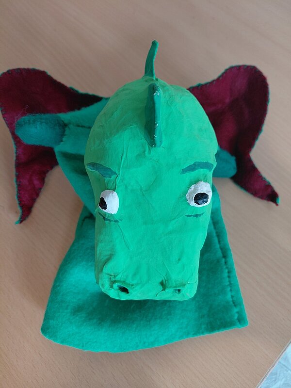 Ein handgefertigter grüner Drache mit roten Ohren sitzt auf einem Tisch und schaut neugierig.