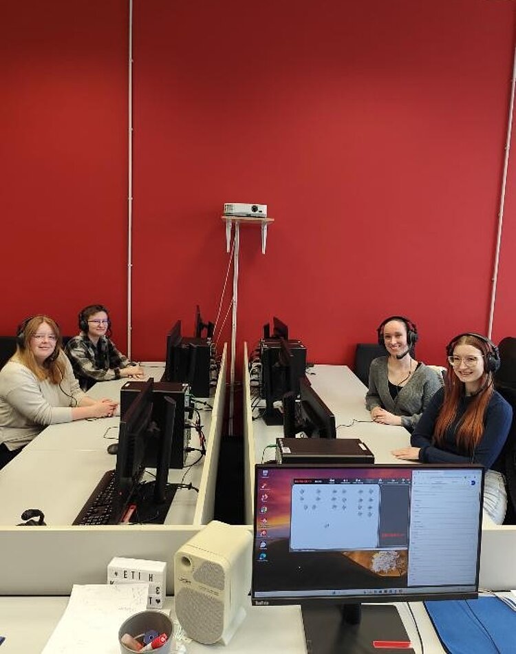 Tag der offenen Tür Vier Absolventinnen der Euro Akademie Bamberg sitzen sich an Tischen, vor dem Computer und mit Headsets auf, gegenüber und posieren für ein Foto.