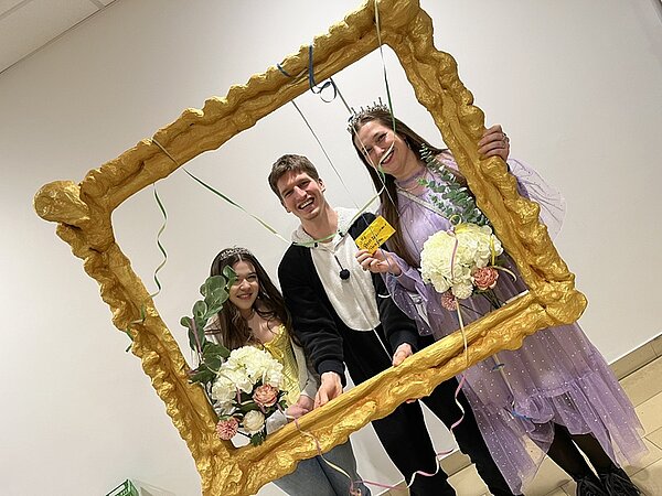 Drei Studierende schauen lächelnd in ihren Faschingskostümen durch einen übergroßen selbst gestalteten Bilderrahmen und posieren für ein Foto.