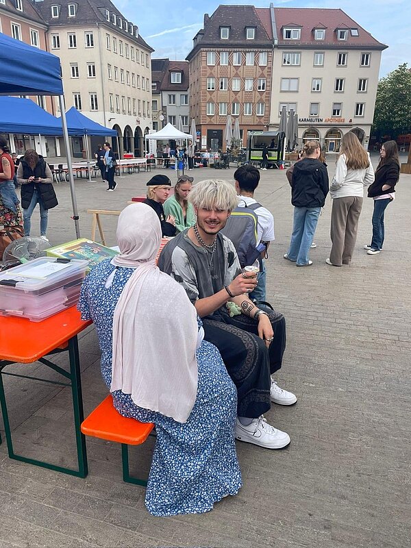 Mehrere Personen sitzen bei einer Veranstaltung auf einem Marktplatz an Biertischgarnituren vor aufgebauten Pavillons.