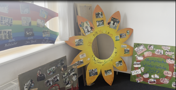 In einem hell erleuchteten Raum stehen mehrere bunte, selbst gestaltete Präsentationstafeln, die anlässlich des Tages der offenen Tür an der Euro Akademie Zwickau entstanden sind. Im Vordergrund befindet sich eine große, gelb-orange Sonnenblume aus Pappe mit einem runden Ausschnitt in der Mitte. Auf den Blütenblättern sind zahlreiche Fotos befestigt, und in der Mitte steht handschriftlich 'Kreis-Land Soziales'. Rechts daneben steht eine grüne Tafel mit der Aufschrift 'Olympische Winterspiele 2017' in blauer Schrift, umgeben von kleinen Bildern und Symbolen, darunter die olympischen Ringe. Links sind weitere bunte Tafeln mit Fotos und Texten zu sehen, die verschiedene Jahre und Themen dokumentieren. Die Tafeln sind auf dem Boden aufgestellt und lehnen an der Wand, die durch ein Fenster Tageslicht hereinlässt.