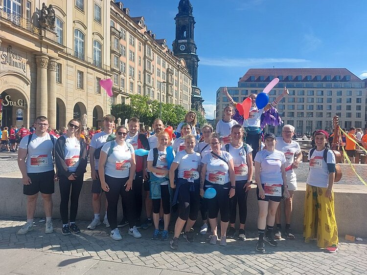 REWE Team Challenge 2024 Eine Gruppe von Männern, Frauen und Kindern in Laufoutfits steht auf einem Platz, im Hintergrund ist ein Kirchturm zu sehen. Einige Personen halten bunte Luftballons in der Hand. Blauer Himmel, die Sonne scheint.