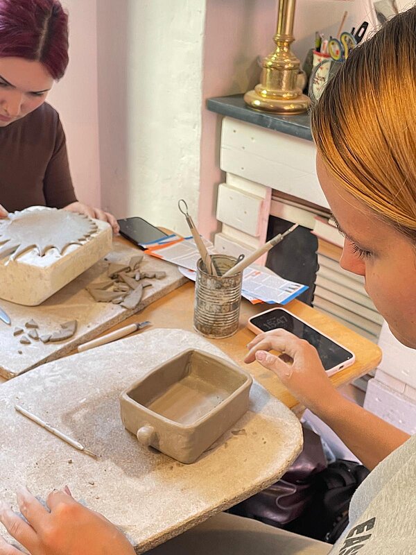 Zwei junge Frauen arbeiten an einem Tisch mit Ton. Im Vordergrund steht eine rechteckige Tonschale, während im Hintergrund eine weitere Tonarbeit mit Modellierwerkzeugen gestaltet wird.