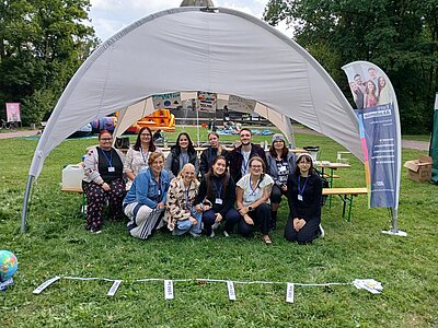 Eine Gruppe von Menschen kniet und steht lächelnd vor einem weißen Zelt auf einer Wiese. Daneben steht ein Banner der Euro Akademie. Im Vordergrund liegt eine Schnur mit Zetteln und daneben steht ein Globus. Im Hintergrund sind bunte Hüpfburgen zu sehen.