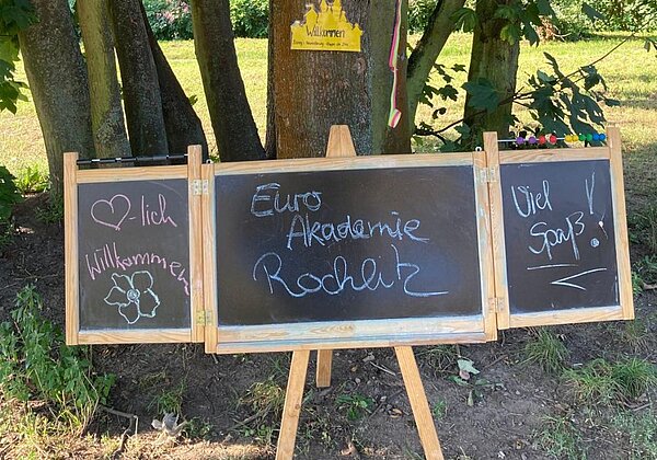 Eine dreiteilige Tafel, auf der "Herzlich Willkommen - Euro Akademie Rochlitz - Viel Spaß!" mit Kreide geschrieben steht, ist mit Hilfe eines Holzgestells draußen vor Bäumen aufgestellt. Die Umgebung sieht wie ein Wald oder Park aus.