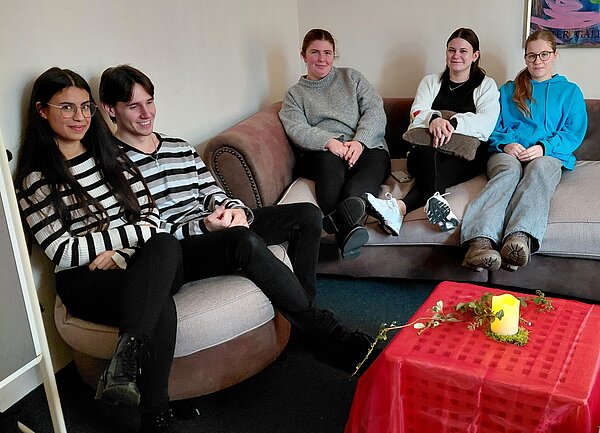 Fünf junge Erwachsene sitzen entspannt auf zwei Sofas, vor ihnen steht ein kleiner Tisch mit rotem Tuch, grüner Deko und einer LED-Kerze, was eine gemütliche Adventsstimmung erzeugt.