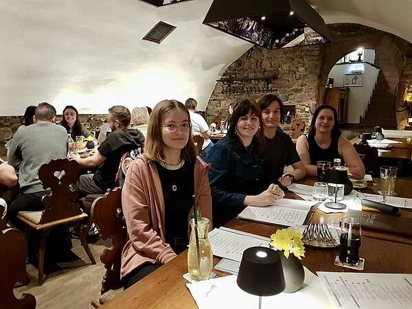 Alumnitreffen 2025 Vier ehemalige Auszubildende der Euro Akademie Aschaffenburg sitzen an einem Tisch im Restaurant und posieren lächelnd für ein Foto. Vor ihnen die Speisekarten, Gläser mit verschiedenen Getränken und eine gelbe Blume in einer kleinen, gläsernen Vase.