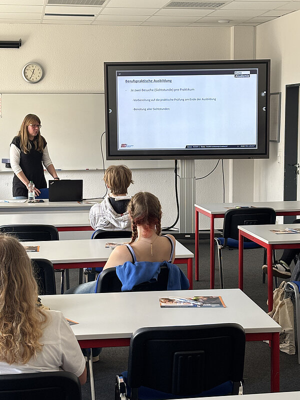 In einem hellen Klassenzimmer lauschen Jugendliche einem Vortrag; auf einer Leinwand ist eine Präsentation zu sehen.