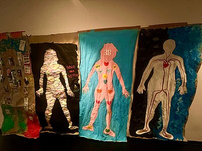 Drei etwas 1 Meter hohe Menschen sind auf jeweils einer Papierleinwand aufgemalt. Die rechte Person ist männlich und auf ihrem Körper sind Adern und ein menschliches Herz aufgemalt, in der Mitte ist eine weiblich Person gemalt mit ganz vielen bunten Herzen auf ihrem Körper und mehreren Plus-Zeichen auf dem Kopf, und ganz links ist ebenfalls eine weibliche Person, deren Körper mit vielen bunten Notizen beklebt ist.