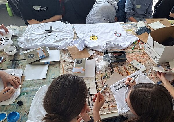 Schülerinnen und Schüler sitzen um einen großen Tisch, auf dem zahlreiche Bastel- und Malutensilien sowie weiße T-Shirts liegen, die sie individuell anmalen und gestalten dürfen.