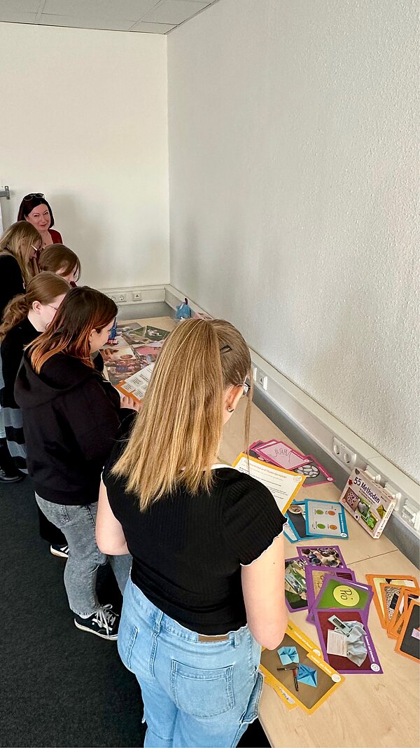 SCHAU REIN! – Woche der offenen Unternehmen Sachsen Mehrere Jugendliche stehen an einem Tisch mit bunten Lern- und Spielmaterialien, darunter Karten, Bilder und Aufgaben – offenbar eine interaktive Lerneinheit.