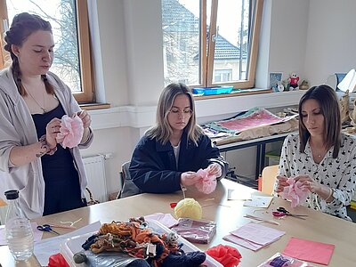 Drei Personen sitzen an einem Tisch und basteln mit rosa Papier und anderen Materialien. Auf dem Tisch liegen verschiedene Bastelutensilien wie Schere, Kleber, Wolle und bunte Papiere.