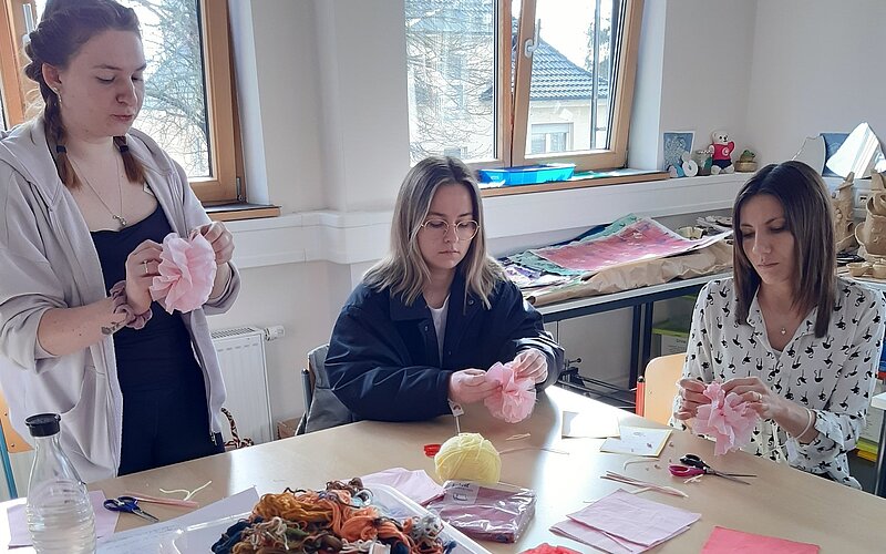 Drei Personen sitzen an einem Tisch und basteln mit rosa Papier und anderen Materialien. Auf dem Tisch liegen verschiedene Bastelutensilien wie Schere, Kleber, Wolle und bunte Papiere.