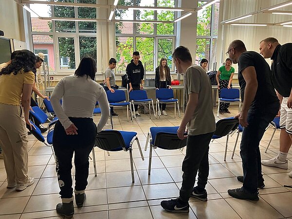 Stuhlkreis mit stehenden neuen Auszubildenden, die ein Kennenlernspiel spielen.