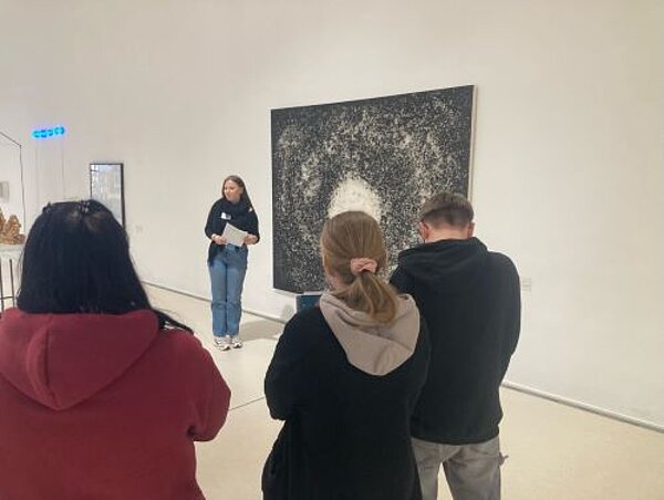 Eine Gruppe von Personen betrachtet ein abstraktes Kunstwerk in einer Galerie, während eine Frau im Hintergrund Informationen dazu vorträgt.
