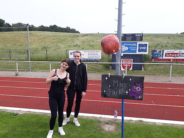Unser Motto zum Sportfest: "Auf zu neuen Galaxien!" Zwei junge Frauen stehen neben einem Schild mit der Aufschrift „Mars Park“. Ein roter Ballon und eine gemalte Alienfigur schmücken das Schild.