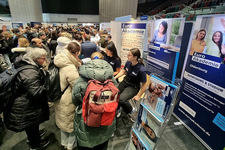 Das Bild zeigt eine Messehalle voller Menschen. Im Vordergrund ist der Messestand der Euro Akademie zu sehen, an dem die Auszubildenden Interessenten beraten.