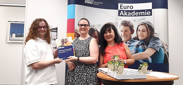 Drei Personen stehen vor einem Roll-up mit dem Logo der Euro Akademie an einem Stehtisch und lächeln. Zwei Personen halten gemeinsam eine Broschüre mit dem Titel "Persönlichkeit durch Bildung". Auf dem Tisch steht eine weiße Palette mit Pflanzen in kleinen Töpfen.