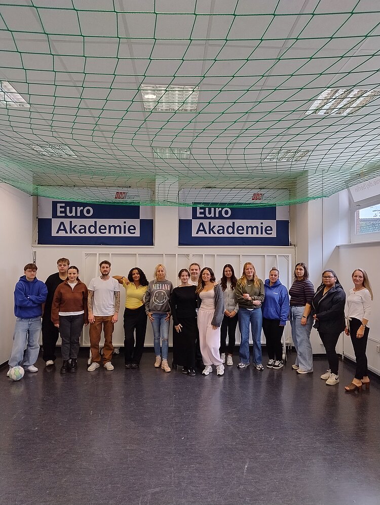 Gruppe von Schülerinnen, Schülern und Lehrkräften steht gemeinsam für ein Gruppenfoto in einem Raum der Euro Akademie. Im Hintergrund sind zwei große Schilder mit dem Schriftzug „Euro Akademie“ sowie ein gespanntes grünes Netz an der Decke zu sehen.