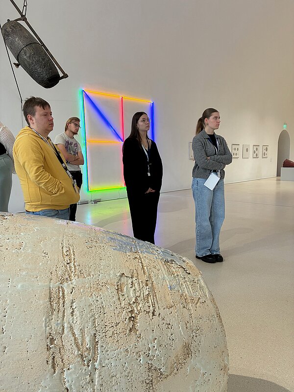 Mehrere Auszubildende zum Erzieher stehen aufmerksam in einem Ausstellungsraum des Kunstmuseums Kloster Unser Lieben Frauen in Magdeburg. Sie betrachten zeitgenössische Kunstwerke im Rahmen einer Exkursion zum Thema frühkindliche ästhetische Bildung. Im Hintergrund leuchten farbige Neonlicht-Installationen in Blau, Rot, Gelb und Grün an einer weißen Museumswand. Rechts sind grafische Arbeiten zu erkennen, links hängt ein großes, dunkles Objekt von der Decke. Im Vordergrund liegt ein helles, strukturreiches Skulpturenelement. Die Lernenden tragen Museumsausweise und wirken konzentriert, während sie die Kunstwerke erkunden. Die Szene vermittelt einen praxisnahen Einblick in kunstpädagogische Bildungsprozesse im Museum.