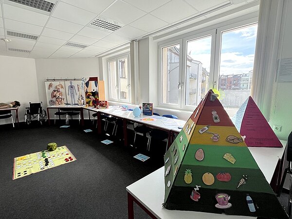 Tag der offenen Tür Im großen Unterrichtsraum sind die Tische an die Wand geschoben, auf ihnen liegen viele unterschiedliche Unterrichtsmaterialien, ein großes Brettspiel auf dem Boden und eine bunte Lehrpyramide auf dem Tisch.