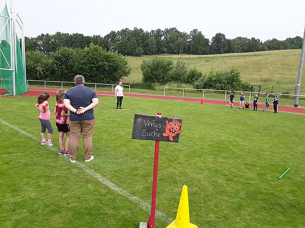 Unser Motto zum Sportfest: "Auf zu neuen Galaxien!" Kinder stehen auf einem Sportplatz in zwei Gruppen, begleitet von einer erwachsenen Person. Im Vordergrund befindet sich ein Schild mit der Aufschrift „Venus Suche“ und einer gezeichneten, roten Alienfigur. Im Hintergrund leitet eine junge Frau das Spiel an. Das Setting ist grün und naturnah mit Laufbahn und Wiese.