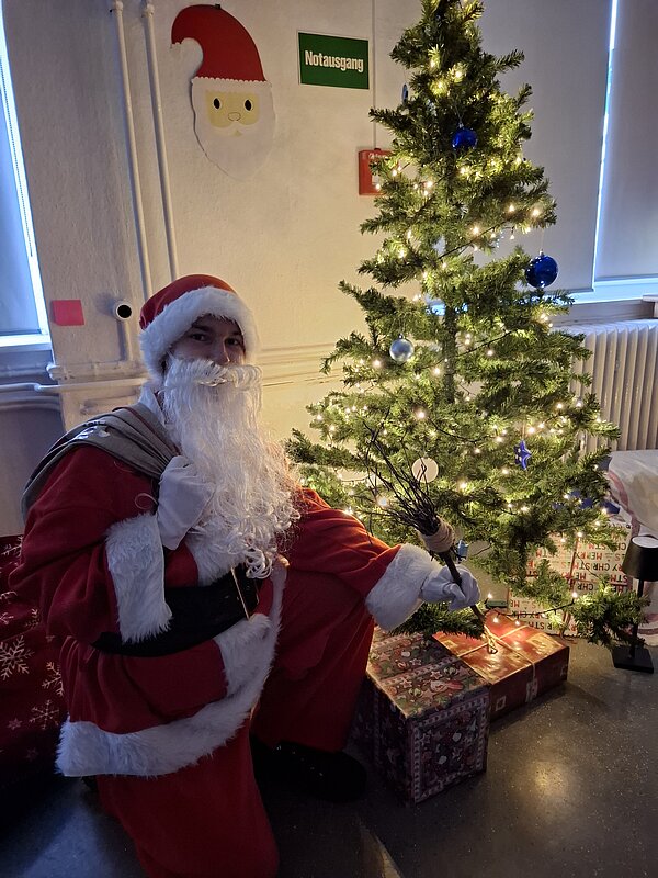 Person im Weihnachtsmannkostüm kniet neben einem geschmückten Weihnachtsbaum mit Lichterkette und Geschenken darunter.