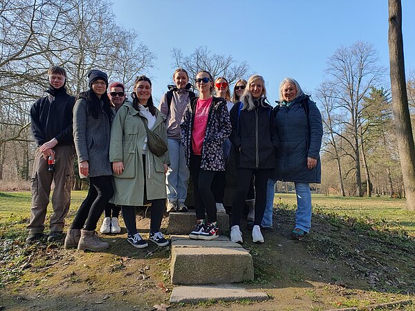 Exkursion in die Natur Eine Gruppe von Menschen steht gut gelaunt auf einer kleinen Steinstufe in einem Park. Es ist sonnig, die Bäume im Hintergrund sind noch kahl. Alle tragen Outdoor-Kleidung.