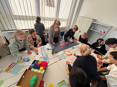 Mehrere Schülerinnen und Schüler arbeiten gemeinsam an einer großen Lernfläche auf dem Tisch. Zwischen ihnen liegen Wollfäden, farbige Karten, Arbeitsblätter und weitere selbst gestaltete Materialien.