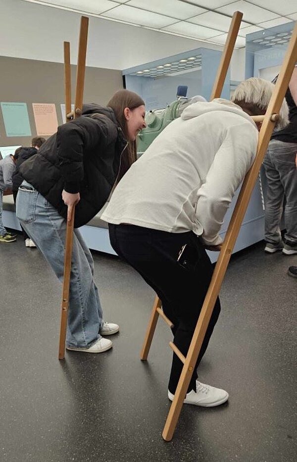 Bewegung und Balance im Hygienemuseum Zwei Personen versuchen auf Stelzen aus Holz zu gehen. Sie beugen sich nach vorne leicht über.