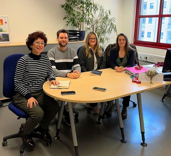 Vier Personen sitzen an einem ovalen Tisch in einem hellen Büro. Sie lächeln in die Kamera. Auf dem Tisch liegen Notizbücher, Handys und einige Papiere. Im Hintergrund sind Pflanzen und ein Fenster mit roten Rahmen zu sehen.