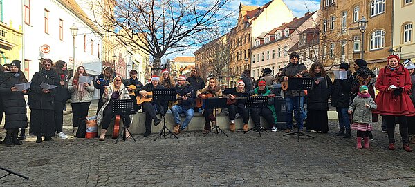 Reihe von Schüler*innen und Lehrkräften, viele mit Nikolausmützen, spielt Gitarre und singt weihnachtliche Lieder auf einem Platz in der Altstadt, umgeben von historischen Häuserfassaden.