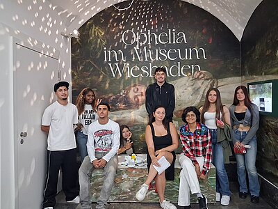 Eine Gruppe junger Schülerinnen und Schüler steht und sitzt vor einer großformatigen Wand mit dem Motiv einer liegenden Frau in einem Gewässer. Die Wand zeigt den Schriftzug 'Ophelia im Museum Wiesbaden'. Die Personen tragen legere Kleidung und blicken in Richtung Kamera. Die Szene spielt in einem Innenraum mit gewölbter Decke, durch die Lichtpunkte auf die Wand und die Personen fallen.