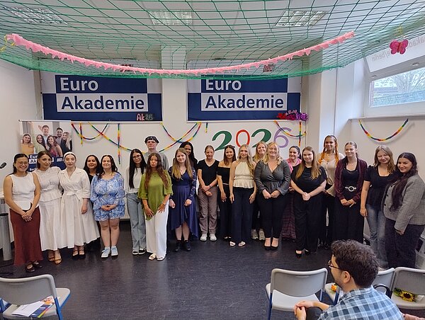 Eine Reihe junger Menschen in festlichen Kleidern steht vor einer Wand mit Girlanden. An der Wand ist in dunkelblau zweimal das Logo der Euro Akademie zu sehen.