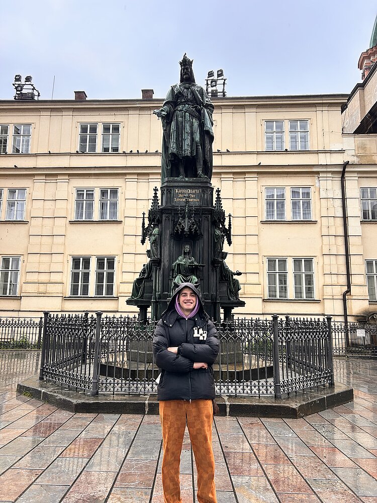 Ein junger Mann steht mit verschränkten Armen steht vor einer großen Statue eines gekrönten Herrschers mit Gefolge auf einem mehrstufigen Sockel, umgeben von schwarzem Gitterzaun. Im Hintergrund zeigt ein gelbes Gebäude mit weißem Putz und grüner Kuppelkuppel ein historisches Setting unter bewölktem Himmel.