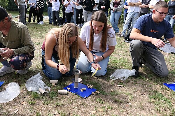 Im Vordergrund knien zwei Schülerinnen auf einer Wiese und beschäftigen sich mit mehreren kleinen Gegenständen, die auf einer blauen Unterlage liegen. Neben ihnen sitzen und knien weitere Schülerinnen und Schüler, die ebenfalls mit ähnlichen Materialien arbeiten. Im Hintergrund stehen mehrere Menschen, die das Geschehen beobachten. Die Szene spielt im Freien, das Gras ist stellenweise abgenutzt. Die Kleidung der Personen ist leger und dem Wetter angepasst.