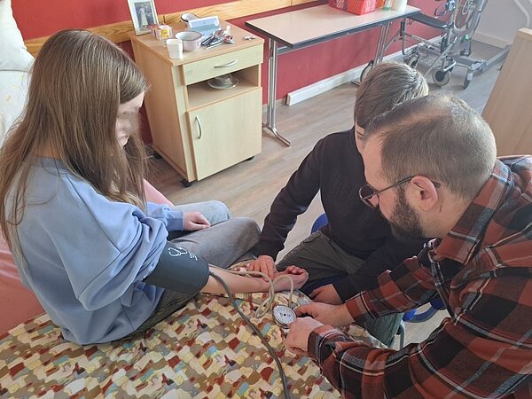 Drei Personen in einem medizinischen Lehrraum: Ein Mädchen sitzt auf einer Liege, während eine erwachsene Person ihr den Blutdruck misst. Eine weitere Person misst mit der eigenen Hand den Puls des Mädchens. Im Hintergrund sind medizinische Geräte und ein Rollstuhl zu sehen.