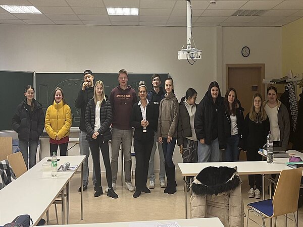 Eine Gruppe von 13 Personen steht in einem Klassenzimmer vor einer Tafel und lächeln in die Kamera. Die Gruppe besteht aus jungen Erwachsenen und zwei älteren Personen, vermutlich eine Lehrkraft und eine Referentin. Die Personen tragen winterliche Kleidung wie Jacken und Pullover. Im Hintergrund sind zwei lange Tische mit Stühlen, eine Tafel mit geometrischen Zeichnungen, ein Projektor an der Decke und eine Tür zu erkennen. Die Atmosphäre wirkt freundlich und aufmerksam.