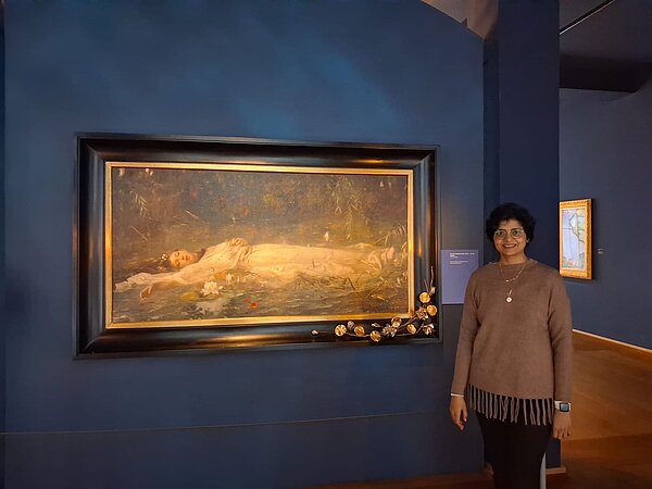 Ein Frau steht neben einem Gemälde von Ophelia an einer dunkelblauen Wand.