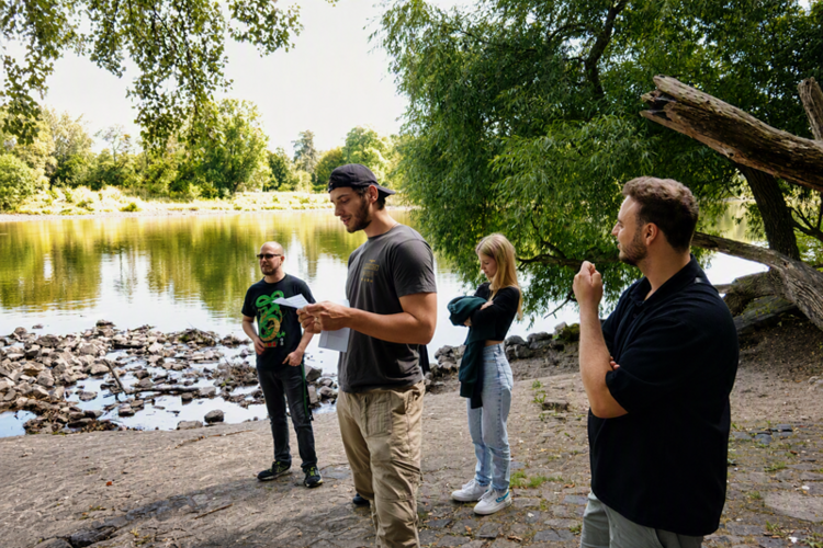 Die Gruppe der Azubis steht auf einem steinigen Ufer vor einem ruhigen See. Im Hintergrund sind Bäume und dichtes Grün zu sehen. Die Personen tragen legere Kleidung; eine Person hält ein Blatt Papier und spricht, während die anderen zuhören. Die Szene wirkt entspannt und findet bei Tageslicht statt.