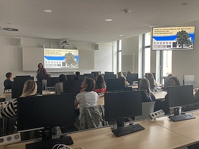 Die Auszubildenden zu Erzieherinnen und Erziehern der Euro Akademie Rochlitz sitzen in einem großen Raum der Universitätsbibliothek Leipzig und hören interessiert der referierenden Mitarbeiterin zu, die vorn eine Präsentation hält.