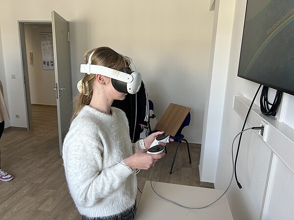 Eine Auszubildende mit hellblondem, zum Zopf gebundenem Haar trägt ein Virtual-Reality-Headset und hält zwei Controller in den Händen. Sie steht in einem hellen Raum mit Holzboden und trägt einen hellen, flauschigen Pullover. Im Hintergrund sind eine offene Tür, ein Stuhl und ein an der Wand montierter Bildschirm zu sehen. Ein Kabel verbindet das Headset mit dem Bildschirm.