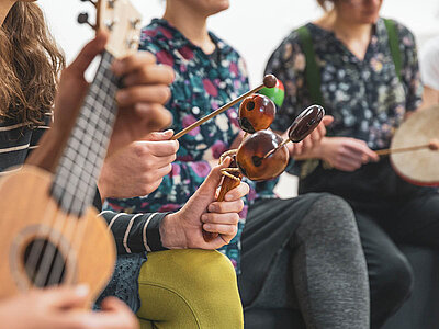 Das Bild zeigt eine Gruppe von Menschen, die verschiedene Musikinstrumente wie Ukulele, Perkussion, Maracas, Rahmentrommel, spielen. Alle Personen der Musikgruppe tragen lässige Kleidung und scheinen sich in einer entspannten Umgebung in einer Freizeitaktivität zu befinden.