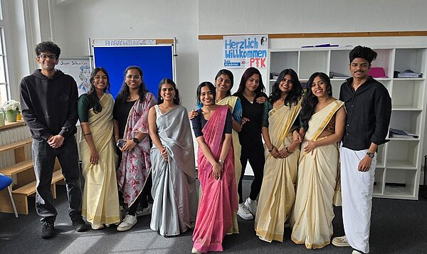 Pflegeklasse feiert Vishu-Fest Eine Gruppe indischer Auszubildender in traditioneller Kleidung haben sich für ein Foto in einem Klassenraum positioniert. Im Hintergrund ist ein Plakat mit der Aufschrift "Herzlich Willkommen PFK" zu sehen.