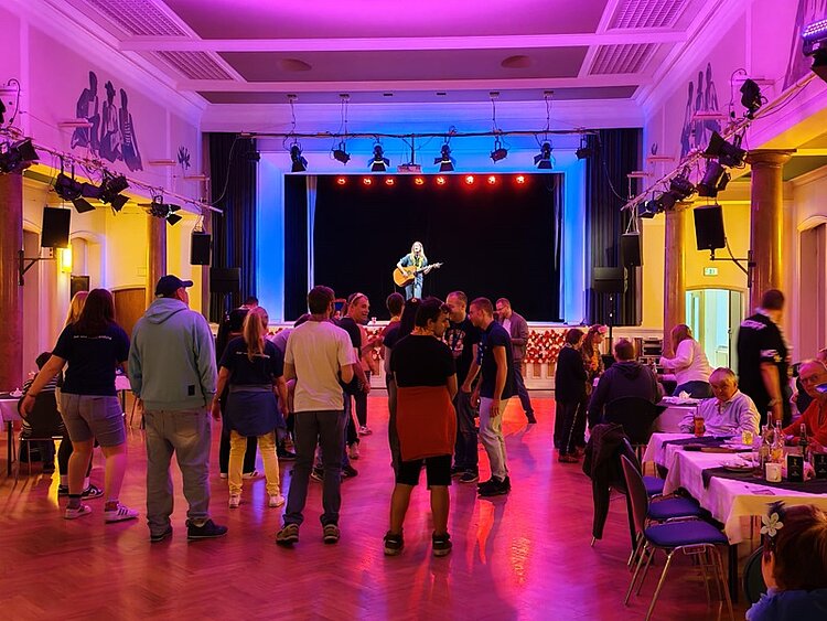 In einem festlich beleuchteten Saal mit hohen Decken und Wandverzierungen steht eine Person mit Gitarre auf einer Bühne im Hintergrund. Der Saal ist in violettes und gelbes Licht getaucht. Im Vordergrund unterhalten sich mehrere Menschen, einige stehen, andere sitzen an gedeckten Tischen am rechten Bildrand. Die Atmosphäre wirkt lebhaft und gesellig.