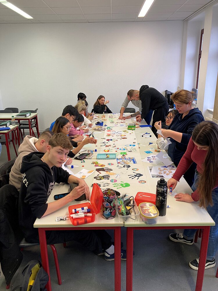 Eingängiges Projekt zum Thema Hygiene Schüler und Schülerinnen sitzen an einem langen Tisch, auf dem ein langes weißes Plakat liegt, auf dem bunte Handabdrücke zu sehen sind. Die Auszubildenden basteln mit Scheren und Kleber. Auf dem Tisch liegen viele bunte Materialien.