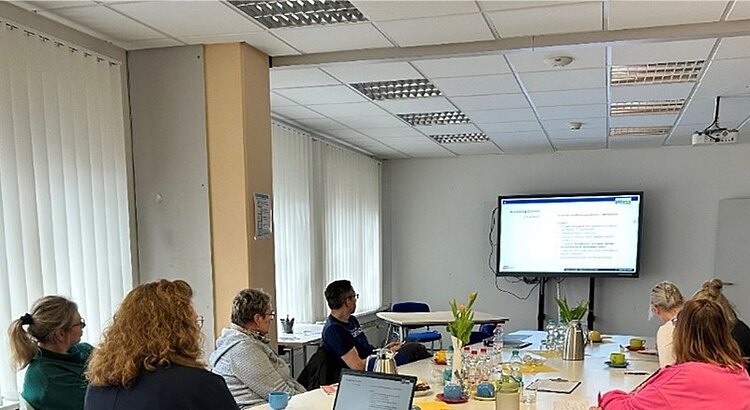 Teilnehmende des Mentorentreffens sitzen in großem Raum am Tisch und schauen nach vorne auf die PowerPoint-Präsentation.